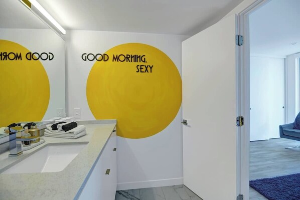 A modern bathroom with a large mirror, a sink, and a towel rack. There are two yellow circular wall decals with text. - Chic Chic Boom- 2 Bedroom Condo, plus office with Full Murphy Bed (Palm Springs)