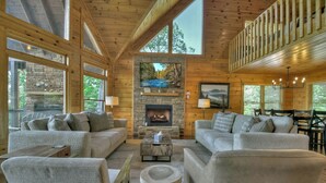 Acogedor interior de una cabaña de madera con chimenea de piedra, ventanas amplias y un balcón.