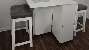 Ett litet vitt bord med två svarta, vadderade pallar, ett vitt skåp och en väggmonterad luftkonditioneringsanläggning.