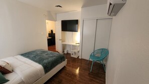A small, well-lit bedroom with a bed, a desk, a chair, and a TV.