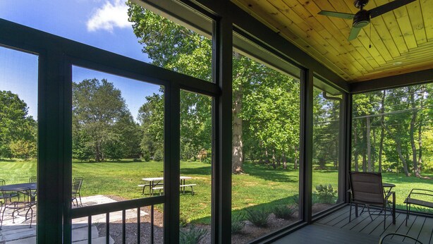 Une terrasse couverte dotée de grandes fenêtres, de mobilier d’extérieur et offrant une vue sur une pelouse et des arbres bien entretenus.