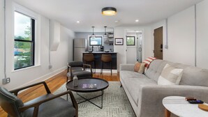 A modern living room with a grey sofa, a round coffee table, and a dining area with two chairs and a kitchen in the background. - Cherry Street Loft (A) | In the Heart of Downtown Black Mtn! (Black Mountain)