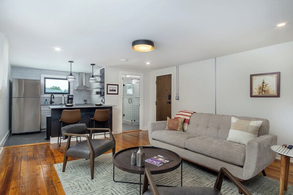 A modern living room with a grey sofa, a coffee table, and a dining area with chairs and a kitchen in the background.