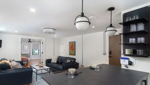 En moderne stue med en mørk sofa, et sofabord og et spiseområde med et stort bord. Der er reoler med glas og en hvid kop på væggen.