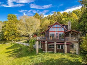 A two-story log cabin with a balcony, surrounded by trees and a clear sky.