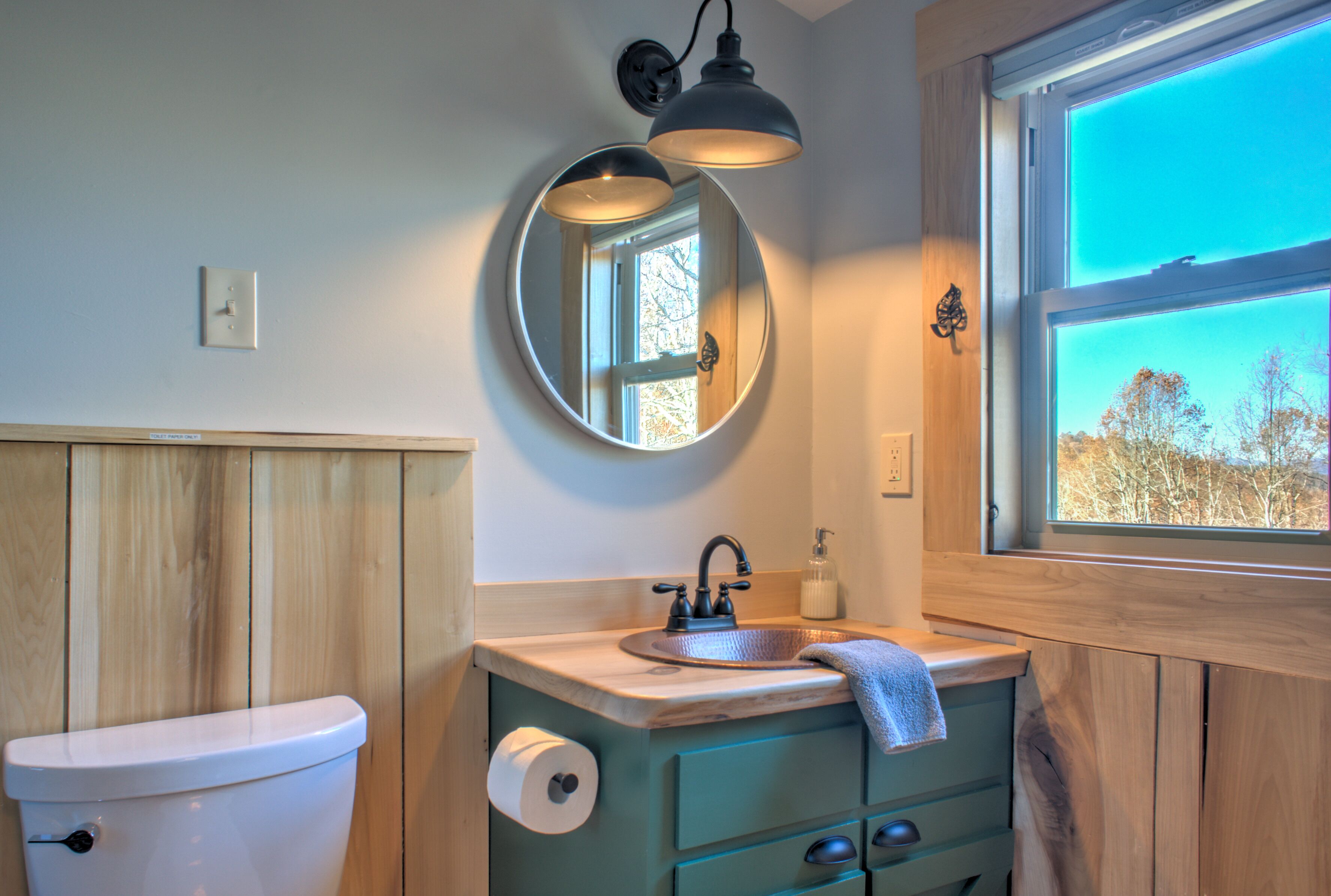A bathroom with a wooden vanity, a round mirror, a sink with a towel, and a window with a view of trees.