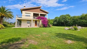 A two-story house with a balcony, surrounded by a well-maintained lawn and flowering plants. - Vivienda Vacacional El Cuetu del Sella (Llames De Pria)