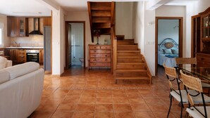 A spacious living area with a wooden staircase leading to a bedroom, a dining area with wicker chairs, and a kitchen with wooden cabinets. - Vivienda Vacacional El Cuetu del Sella (Llames De Pria)