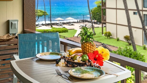A table with plates, a pineapple, and tropical fruits on a balcony overlooking a beach. - 🏖 ️Pretty Ocean View | Top Floor | Concierge Included (Kailua Kona)