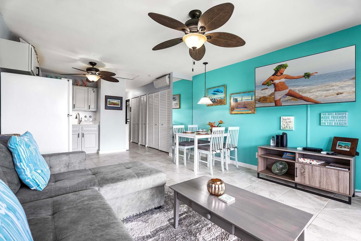 A modern living room with a grey sofa, a coffee table, and a dining area with white chairs. There are ceiling fans, a kitchen area, and a large beach-themed painting on the wall.