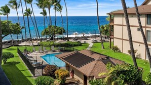 A pool area with a view of the ocean, surrounded by palm trees and a building with a tiled roof. - 🏖️Pretty Ocean View | Top Floor | Concierge Included (Kailua Kona)
