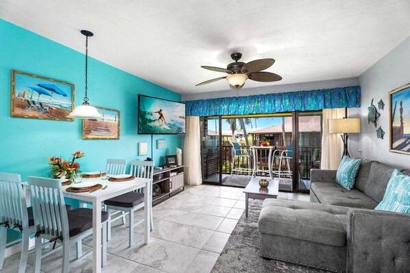 A bright living room with a teal wall, a ceiling fan, a dining area with white chairs, and a grey sofa. - 🏖️Pretty Ocean View | Top Floor | Concierge Included (Kailua Kona)