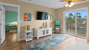 Sala de estar com televisão, obras de arte, ventilador de teto e vista para um deck externo.