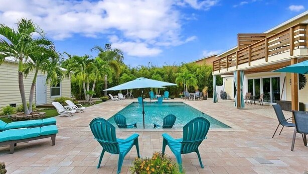Piscina com espreguiçadeiras e guarda-sóis, cercada por palmeiras e um espaço coberto com varanda.