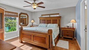 Een slaapkamer met een houten bed, twee nachtkastjes met lampen, een plafondventilator en een raam met uitzicht op groen.