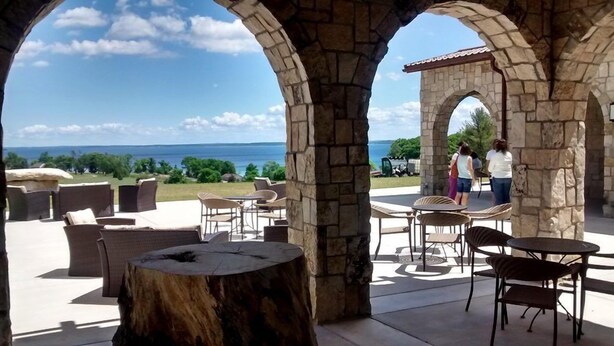 A stone-arched outdoor seating area with wicker furniture and a round table, overlooking a scenic view of the sea. - 539 Bay St. Lake View Condo Unit 203E (Traverse City)