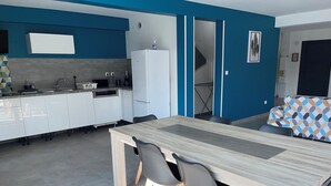 Una cuina moderna amb armaris blancs, forn integrat i nevera blanca. Hi ha una zona de menjador amb taula de fusta i quatre cadires.