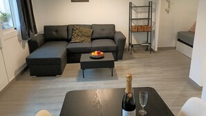 Un salon moderne avec un canapé d’angle, une petite table sur laquelle se trouve un panier de fruits, une bouteille de champagne et un verre. On y trouve également une bibliothèque avec des paniers et une fenêtre avec des rideaux.