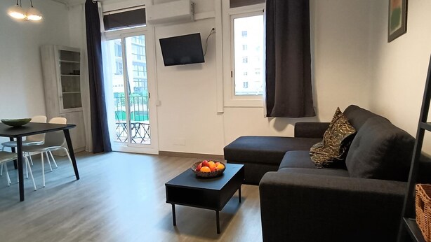 Un salon moderne avec un canapé, une petite table sur laquelle se trouve un panier de fruits, un tél éviseur fixé au mur et un balcon visible à travers la porte.