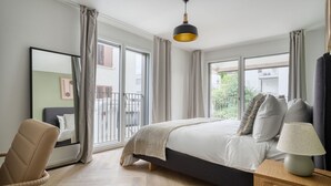 Un dormitorio con cama, una silla, una mesita de noche con lámpara y una ventana con cortinas.