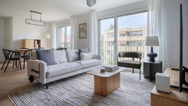 Una sala de estar moderna con un sofá, una mesa de centro y sillas. Hay un comedor con una mesa y sillas. Amplias ventanas ofrecen vistas al exterior.