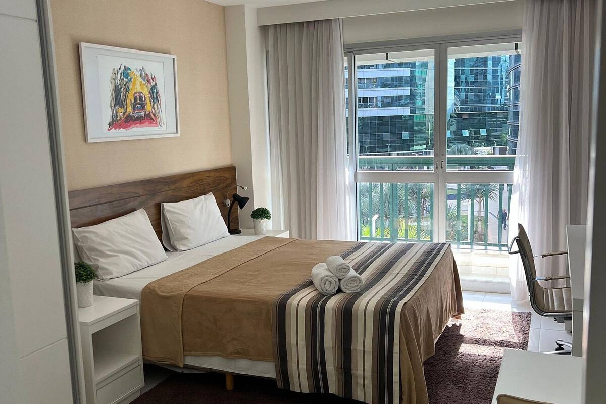 Een hotelkamer met een bed, nachtkastjes, een bureau en uitzicht op de stad door grote ramen.