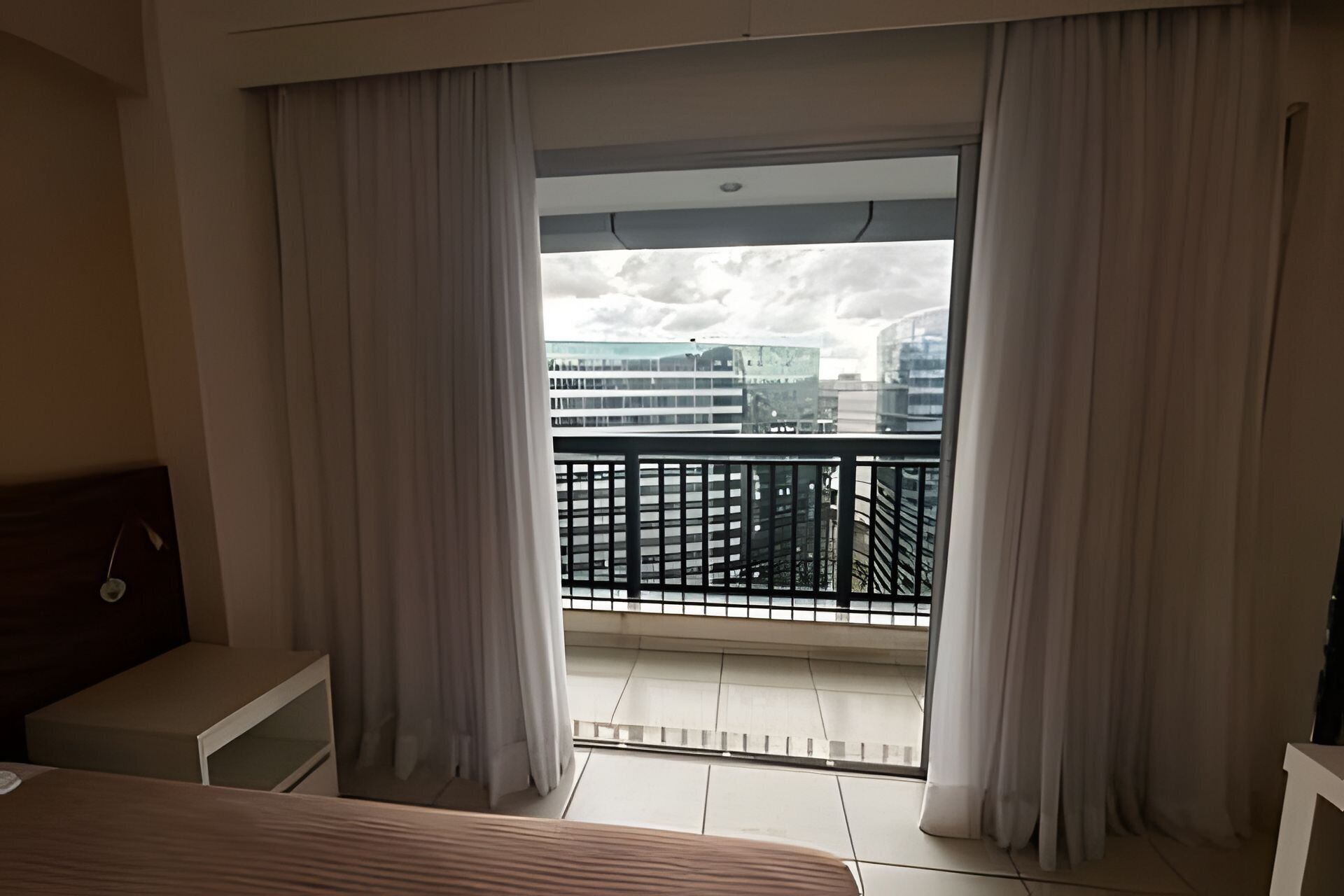 A hotel room with a balcony, a bed, a nightstand, and a view of city buildings.