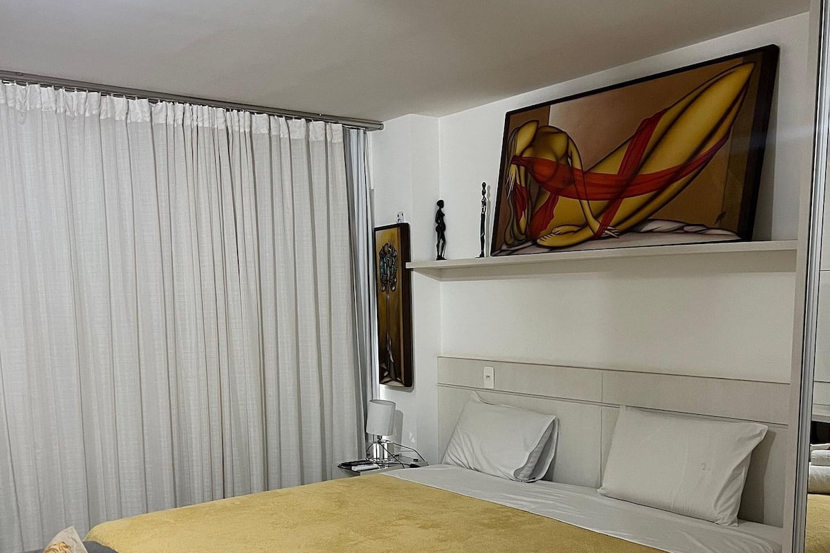 Een hotelkamer met een bed, een grijze fauteuil, een gele deken en een schilderij aan de muur.