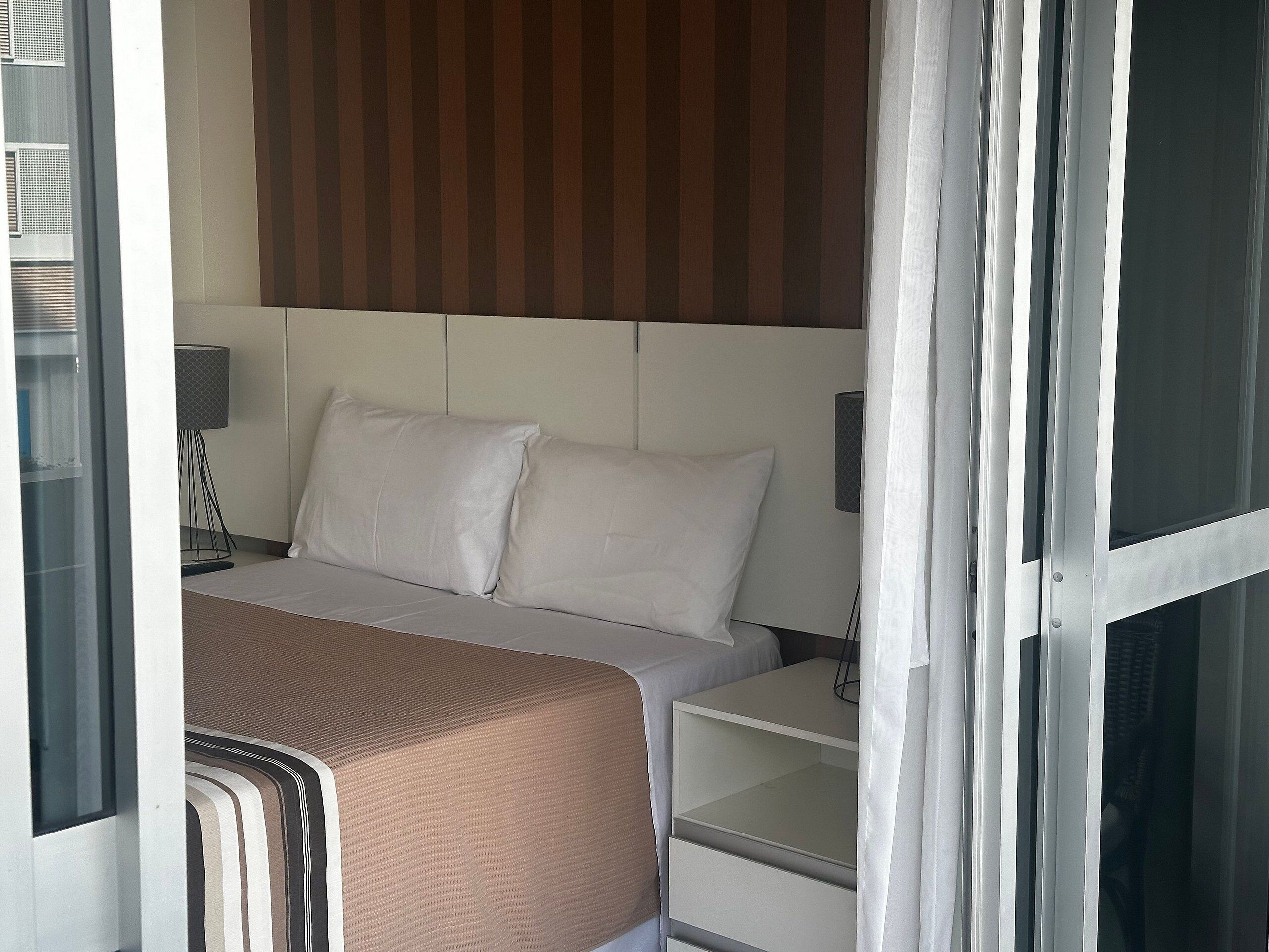Quarto de hotel com cama, mesa de cabeceira, luminária e janela com cortinas.