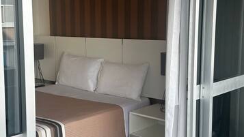 Quarto de hotel com cama, mesa de cabeceira, luminária e janela com cortinas.
