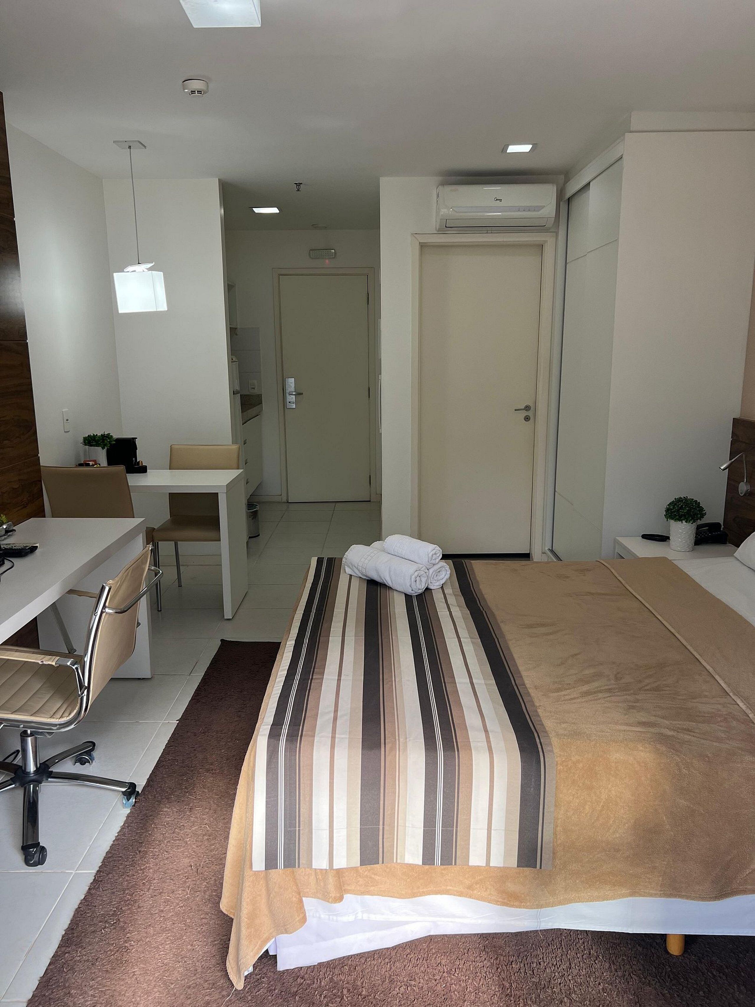 A hotel room with a bed, a desk, a chair, a small table, and an air conditioning unit.