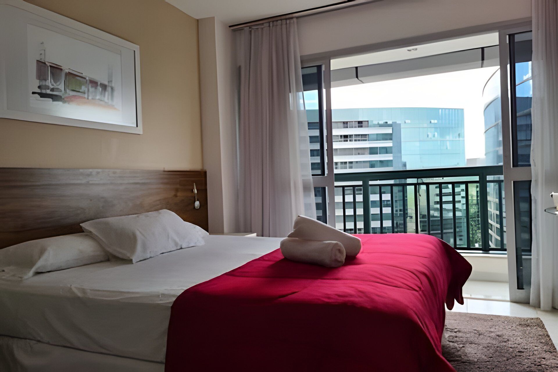 A hotel room with a bed, a red blanket, a white pillow, a framed picture on the wall, and a balcony with a view of buildings.