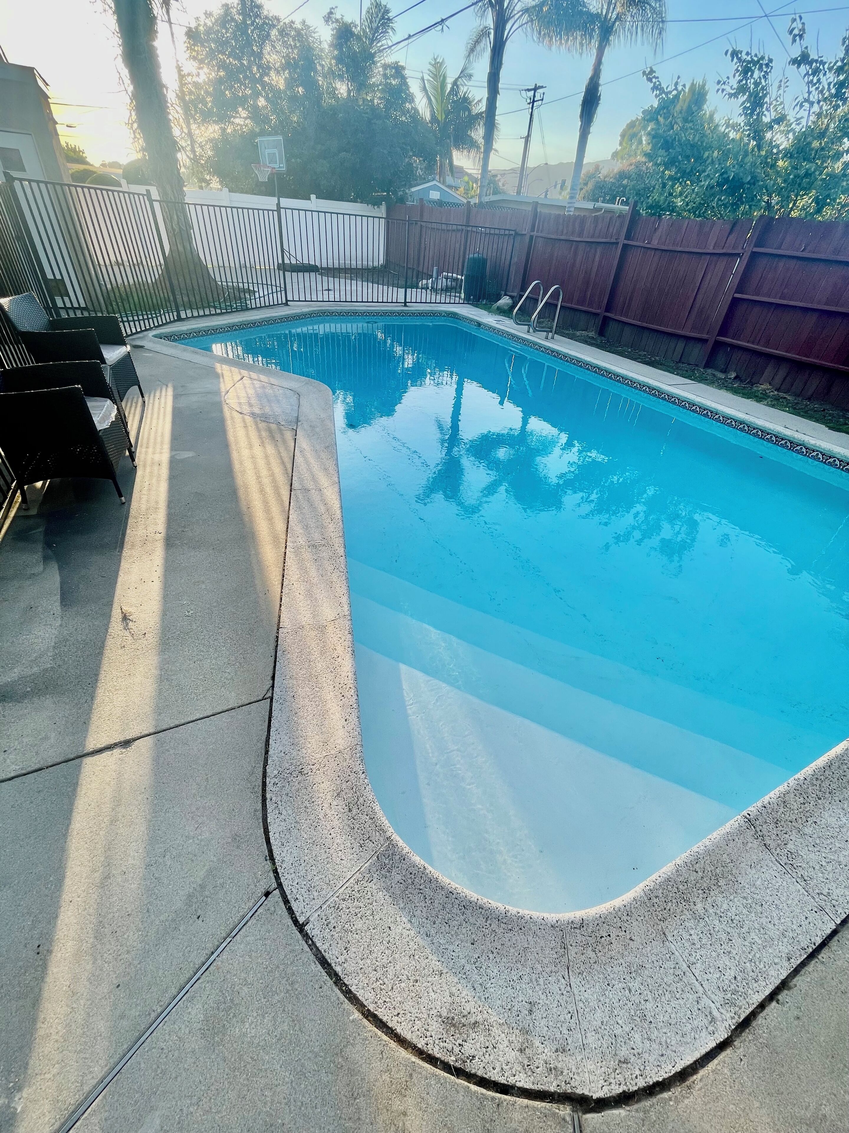 Ein rechteckiger Swimmingpool mit Betonrand, umgeben von einem Holzzaun und einem Metalltor.