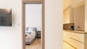 A modern apartment with a wooden floor, a bed with a gray headboard, a small bedside table, and a kitchenette with a coffee maker. - Serenity & Luxury - Elegant 2BR in Kifisia's Center (Kifisia)