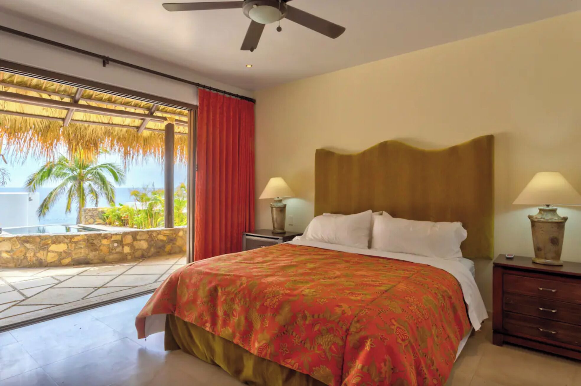 A hotel room with a bed, two bedside lamps, a wooden headboard, a window with a view of palm trees and a balcony, and a ceiling fan.