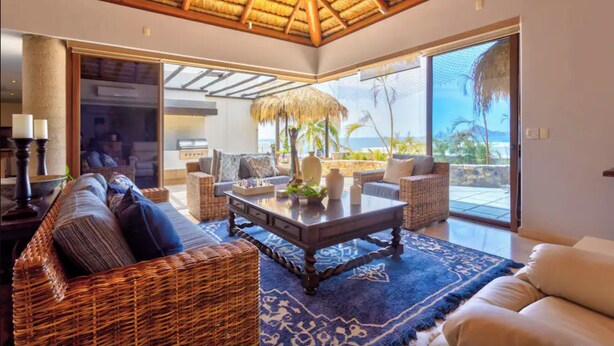 A spacious living area with a thatched ceiling, wicker furniture, and a large window overlooking a scenic view. - Infiniti Pool-View of Arch, Oceanview-Walk to Beach, Cape Hotel, Close to Dwntn  (Cabo San Lucas)