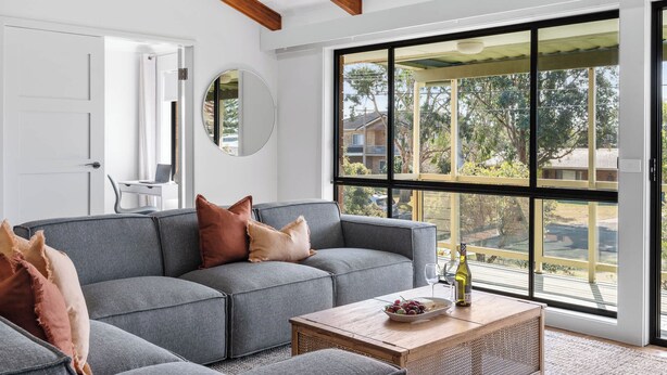 Una sala moderna con un sofá seccional gris, una mesa de centro de madera y ventanas amplias que ofrecen vistas al exterior.