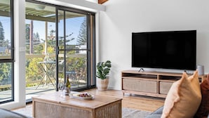 Una sala de estar moderna con un televisor grande, una mesa de comedor con una botella y copas, y un balcón con mesa y sillas.