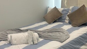 Ein ordentlich gemachtes Bett mit gestreifter Bettwäsche und zwei Kissen, ein zusammengerolltes Handtuch und ein Fenster mit Vorhängen.