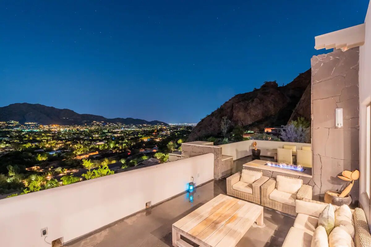 A rooftop terrace with a view of a cityscape at night, outdoor seating, and a fire feature.
