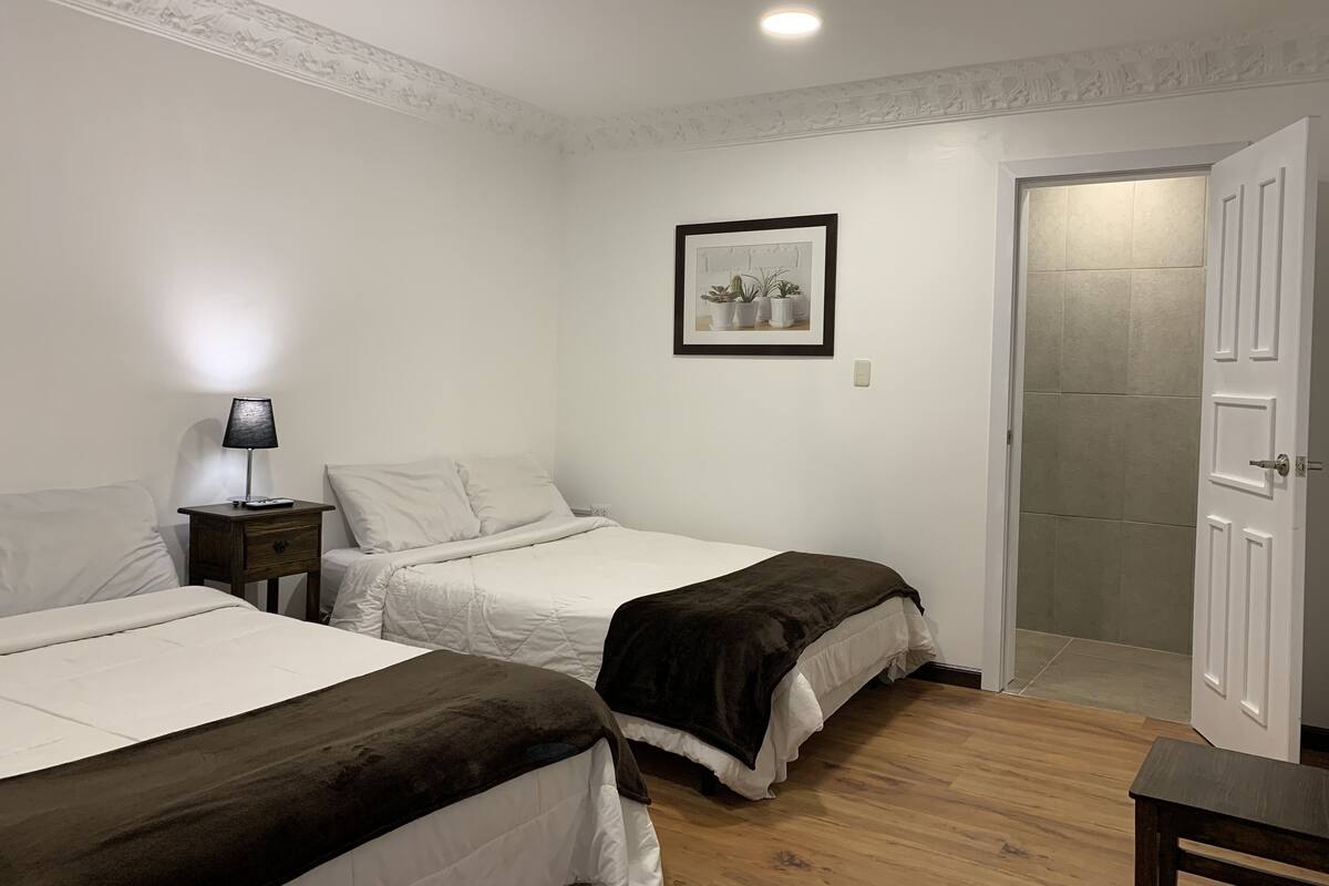 A bedroom with two beds, a nightstand, a lamp, a framed picture, and a door leading to a bathroom.