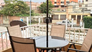 Mesa redonda con cuatro sillas y un paraguas grande en un balcón con vistas a edificios y vegetación.