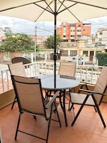 Mesa redonda con cuatro sillas y un paraguas grande en un balcón con vistas a edificios y vegetación.