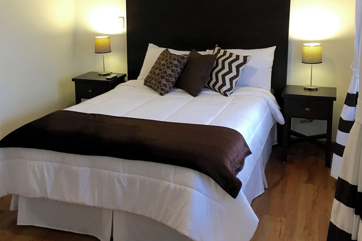 A neatly made bed with a dark headboard, white bedding, and decorative pillows. There are two bedside tables with lamps, and a striped curtain is partially visible.