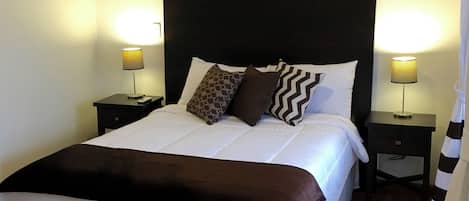 Una cama cuidadosamente hecha con un cabecero oscuro, ropa de cama blanca y almohadas decorativas. Hay dos mesitas de noche con lámparas, y se ve parcialmente una cortina a rayas.