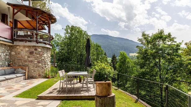 Un edificio de piedra con una terraza de madera, mobiliario exterior y vistas a montañas.