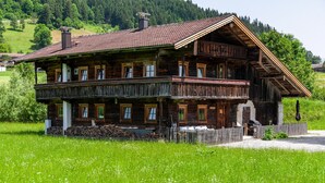 A wooden multi-story house with balconies, surrounded by a grassy field and a forested hill. - Holiday Home "The Farm" with Mountain Views, Private Terrace & Wi-Fi (Westendorf)