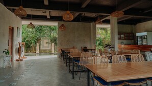 A spacious dining area with wooden tables and wicker chairs, large windows offering a view of greenery, and hanging wicker lamps.