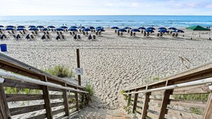 Ein Strand mit Reihen blauer Sonnenschirme und Liegestühle, Holzstege und freier Sicht auf den Ozean.