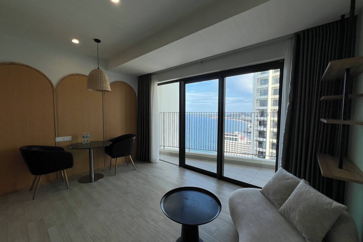 A modern living room with a sofa, a dining table, and chairs. There is a large window with a view of the city and a balcony.